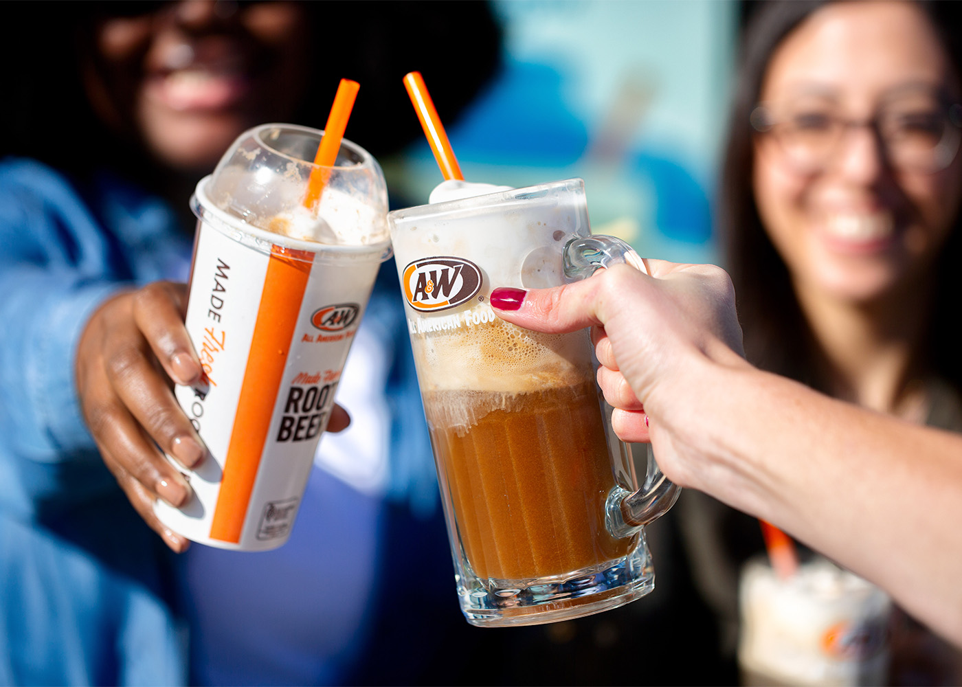 Two people outdoors toasting. One person is holding an A&W Root Beer Float in a to-go cup. The other person is holding an A&W Root Beer Float in a frosty mug.